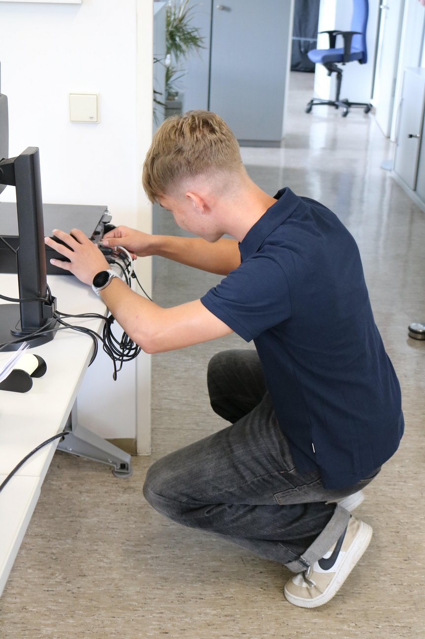 Junger Mann kniet vor einem Schreibtisch und steckt Kabel in eine Docking Station