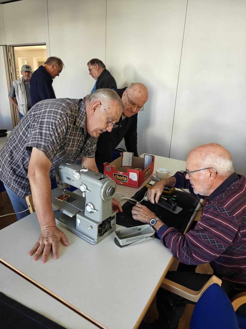 Reparaturwerkstatt_2.jpg GDrei Männer beugen sich über eine reparaturbedürftige Nähmaschine und reparieren diese.